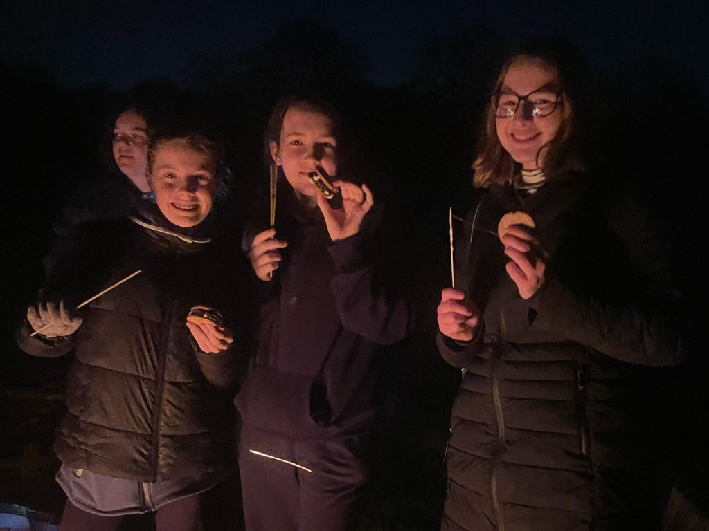 Three girls in winter coats standing in the dark holding sticks and eating smores.