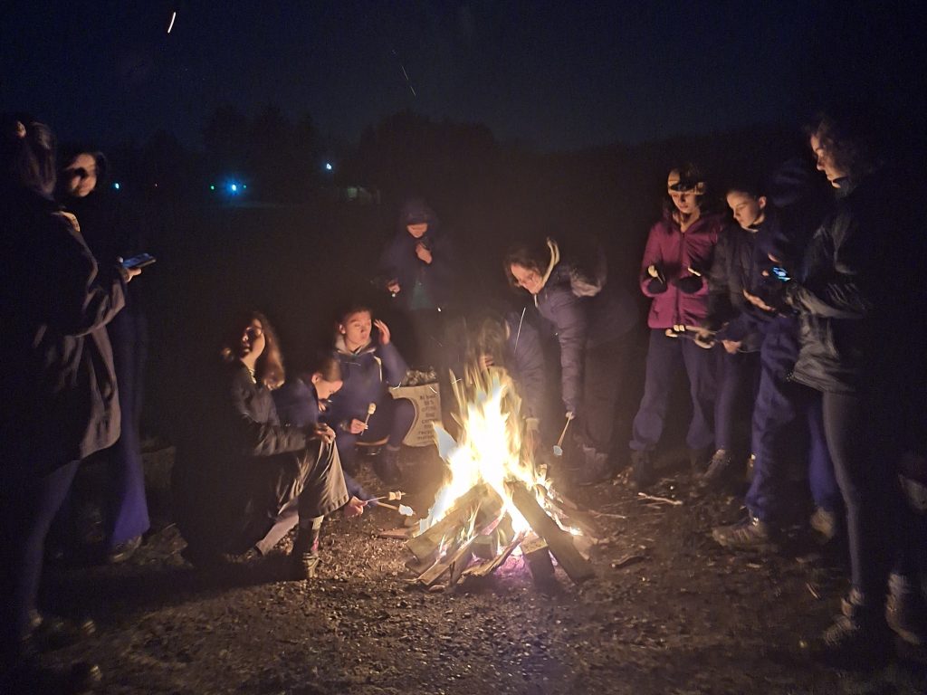 Nighttime. Girls in coats stand around a camp fire toasting marshmallows on sticks.