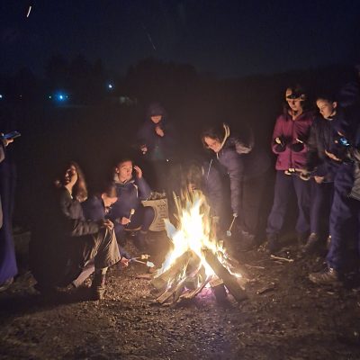 Nighttime. Girls in coats stand around a camp fire toasting marshmallows on sticks.