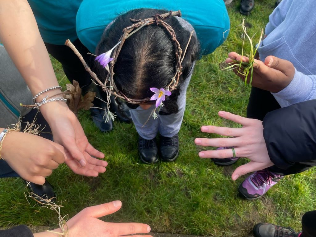 Girls showing jewellery made from nature. Girl in centre has twig crown with purple crocus flowers, others have grass rings and bracelets made from straw and autumn leaves.