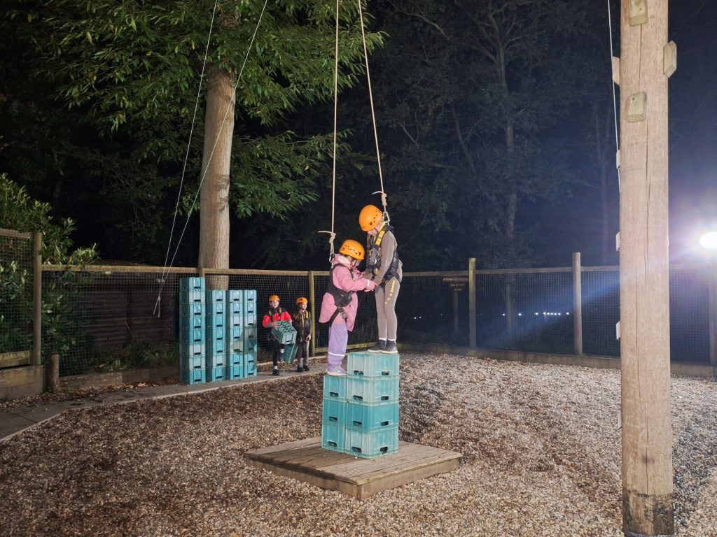 Under a dark sky, two brownies in yellow safety helmets help each other as they stand on a stack of blue plastic crates. Two other girls stand in the background next to a large pile of crates.