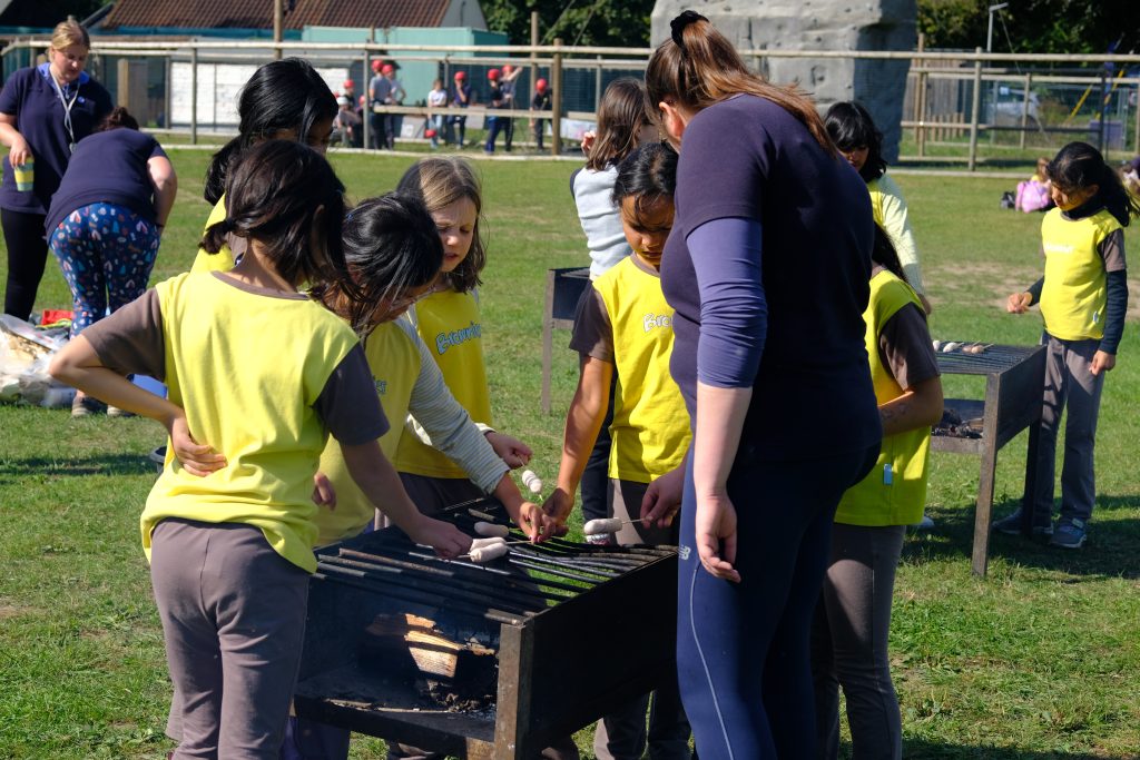 Girls gathered around a metal barbecue with their leader. They're cooking sausages on sticks.