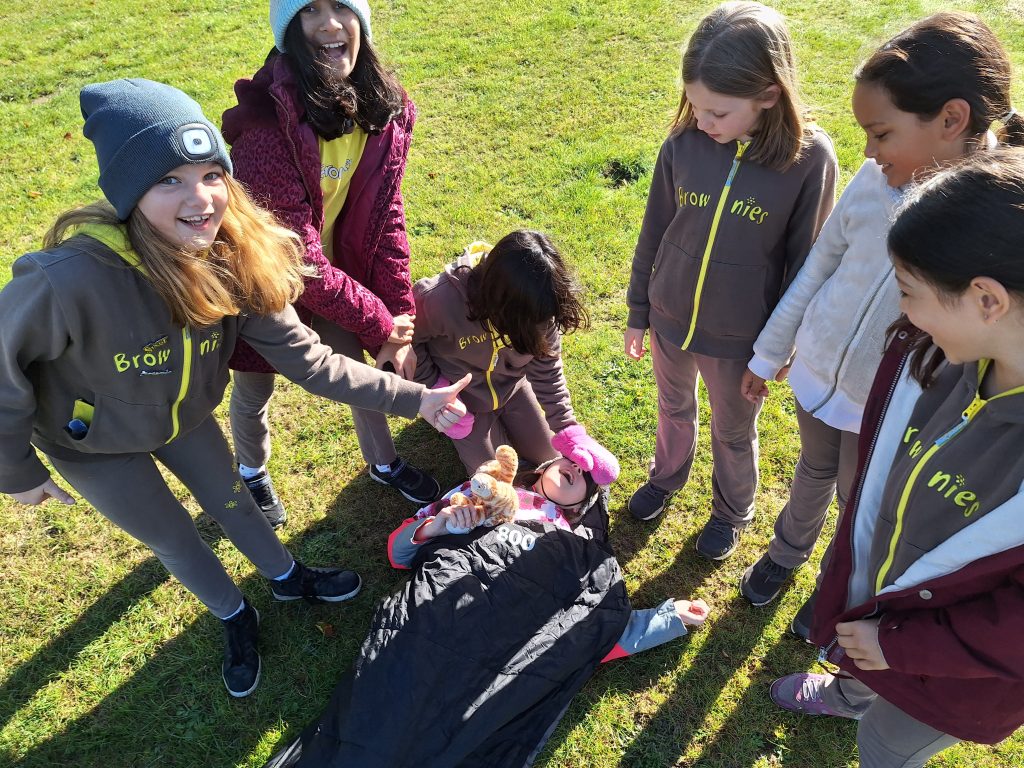 A girl lying on the ground in her sleeping bag as her eyes are covered by a girl. Andother girl grins and gives a thumbs up.