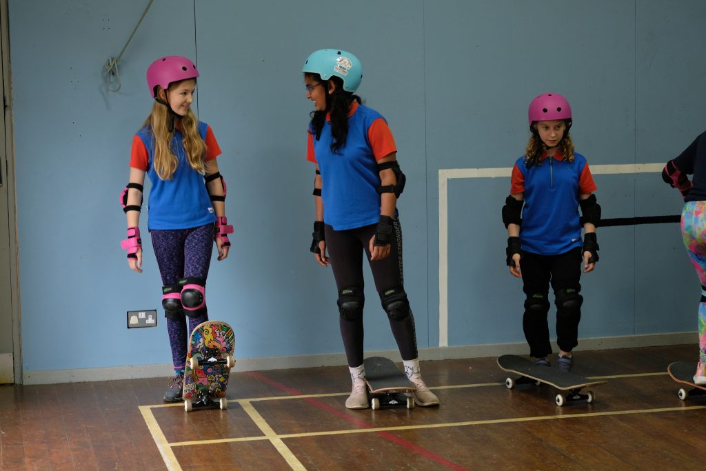 Two Guides in safety helmets grin at each other as they stand with their skateboards. Another girl stands nearby.