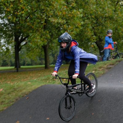 A Guide cycles over a hump on the tree-lined track, her blue coat flying out behind her as she goes. Another girl waits with her bike in the background.
