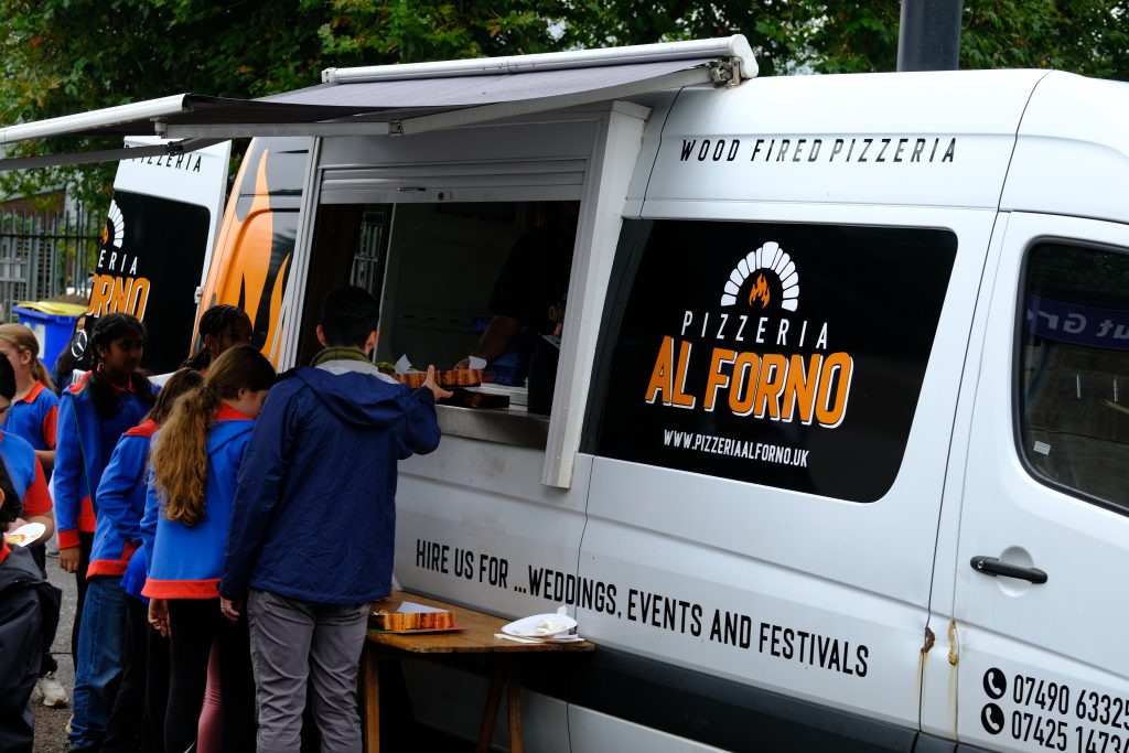 People waiting by a white pizza truck with an awning over the hatch. The logo, Pizzeria Al Forno is on the side of the van.