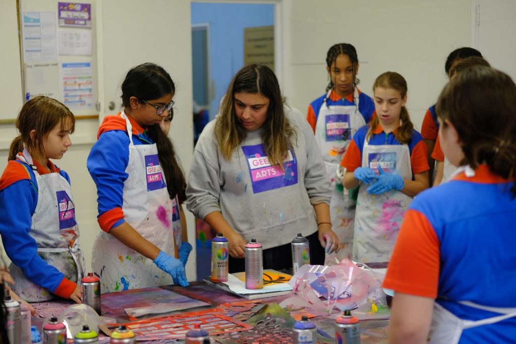 Guides in their uniforms and wearing aprons and gloves stand with an instructor as they do spray painting at a table.
