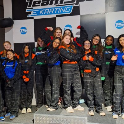 A group of girls in their karting jumpsuits standing on and around the winners' podium. Two in the centre stand with their arms folded and with their backs to each other. Two behind flex their muscles.