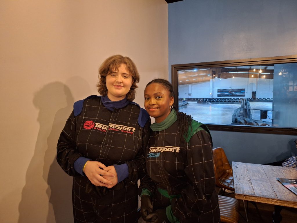 Two Rangers, one white with short blonde hair, and the other black with long dark hair. They're smiling as they stand in front of a viewing window looking out onto the karting track.