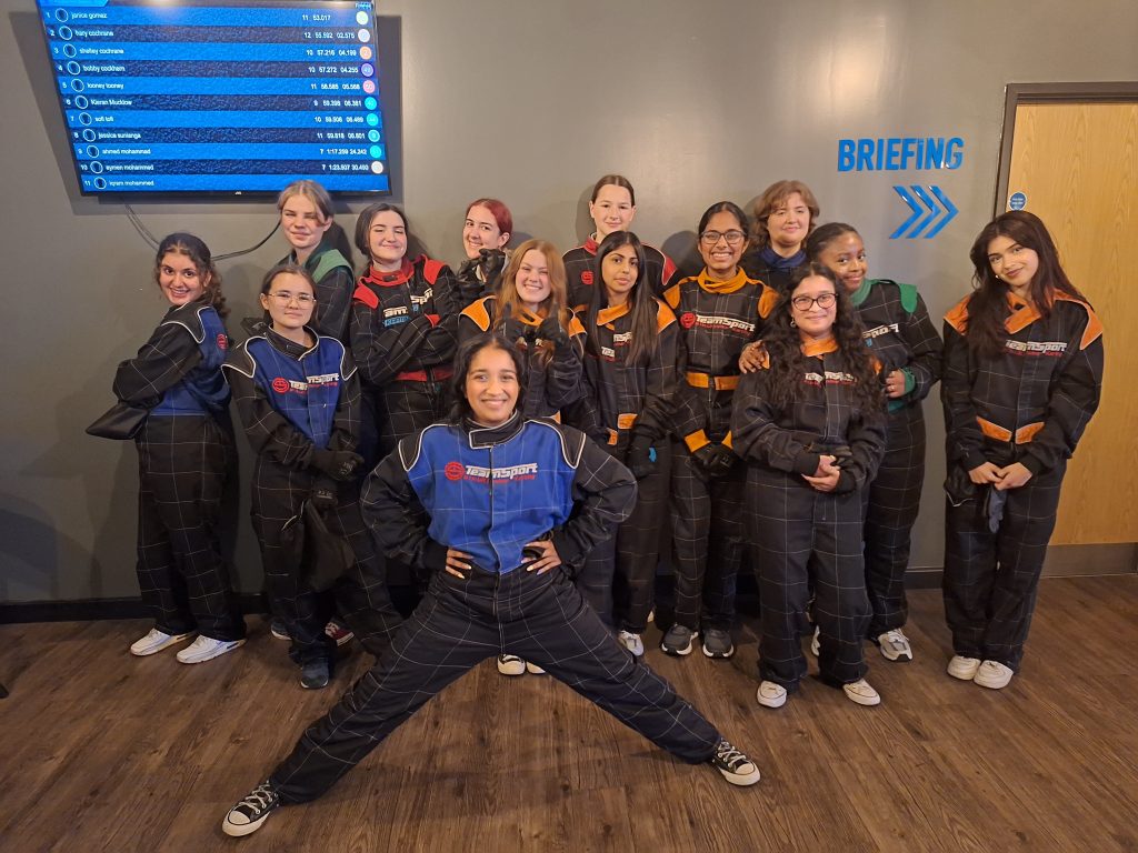 A group of girls in their karting jumpsuits. One stands in front with her legs stretched to the side and with her hands on her hips. They're in front of the Race Position monitor.