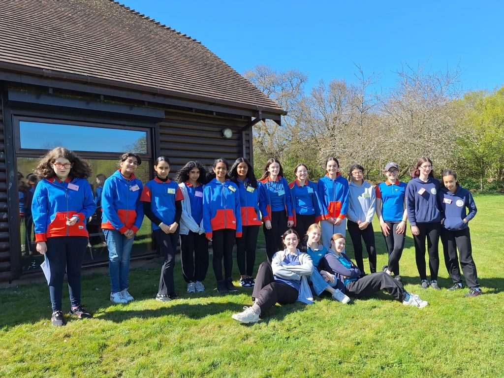 A large group of Guides standing outside a building on a sunny day with three Rangers sitting on the grass in front of them. There are trees in blossom in the background.