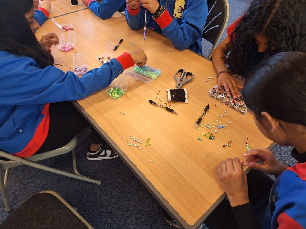 A view of the table with containers of colourful beads, black elastic and scissors. The girls are looking through the beads and threading them onto elastic.