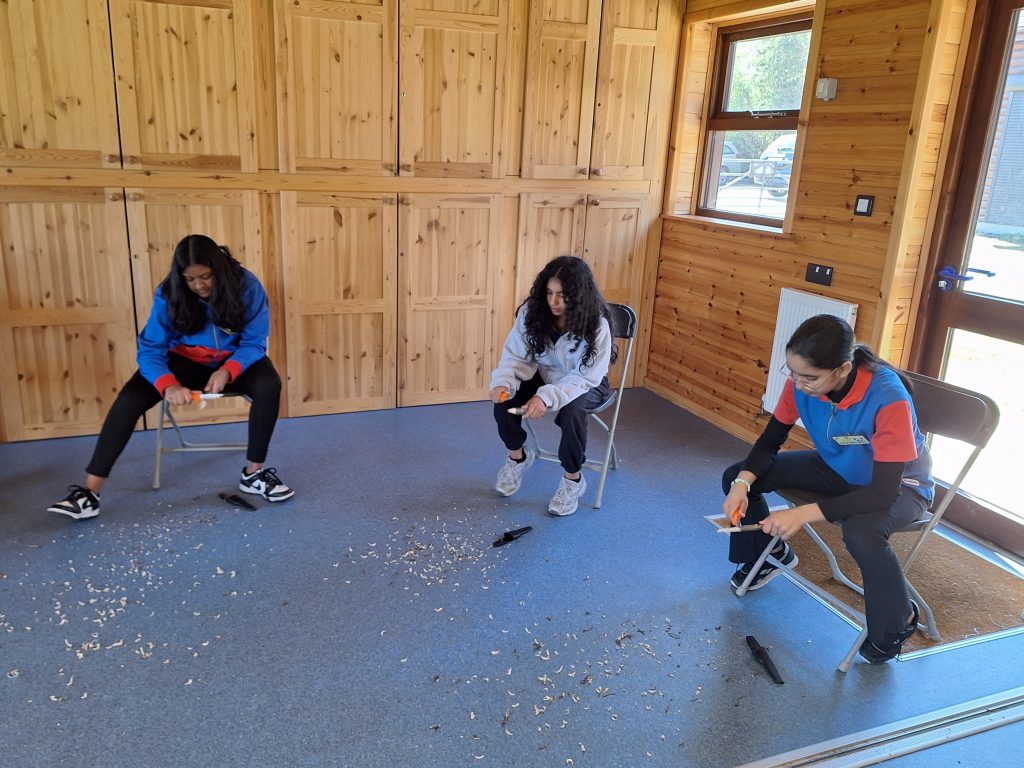 Three girls sitting in a wood panelled room, leaning over as they whittle. Wood shaving litter the floor in front of them.