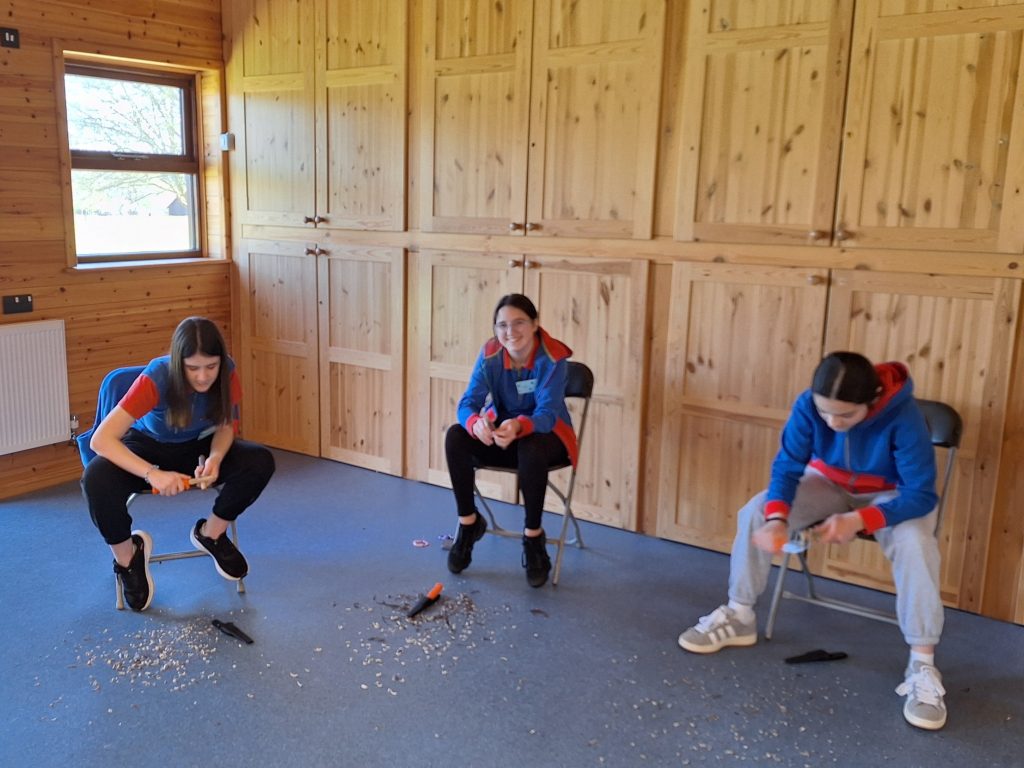 Three Guides sitting in a wood panelled room, whittling. The girl in the centre is looking up and smiling.